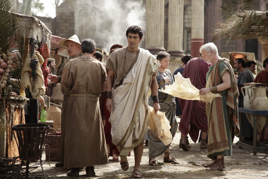 古代ローマの市場で人々が活動している様子。トーガを着た男性が道を歩き、他の人々が商品を扱っている。