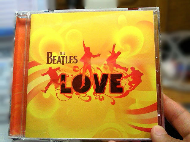 ビートルズのアルバム『LOVE』のCDジャケットを手に持つ人物の手元。ジャケットはオレンジと黄色のデザインで、中央に大きな黒い文字で「LOVE」と書かれている。周囲にはシルエットのダンスをする人々のイラストが描かれている。