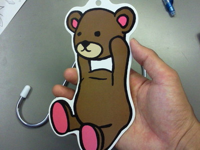 手に持たれた茶色のクマのキャラクターが描かれた飾り。背景にはS字フックがあり、手前には手が見える。