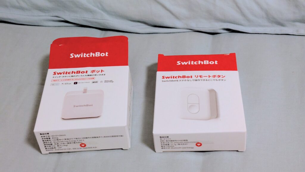 SwitchBotのボックスとリモートボタンのパッケージがベッドの上に置かれている画像